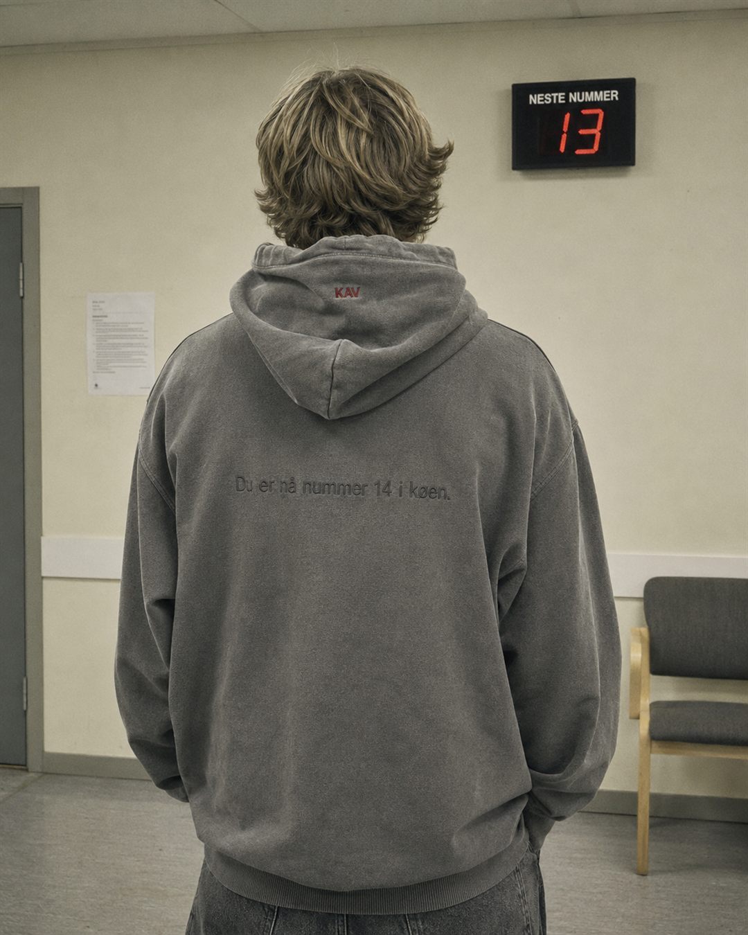 Modell bakfra i grå hettegenser med brodert «Du er nå nummer 14 i køen» foran kønummerdisplayet